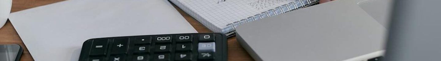 A woman writes financial calculations in a notebook, using a calculator and laptop at a wooden desk.