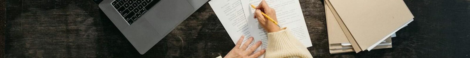 Overhead view of woman writing at desk with laptop, papers, and books.