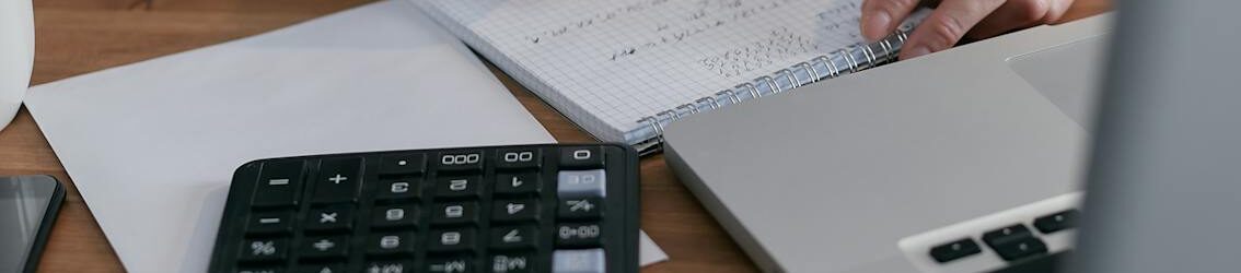 A woman writes financial calculations in a notebook, using a calculator and laptop at a wooden desk.