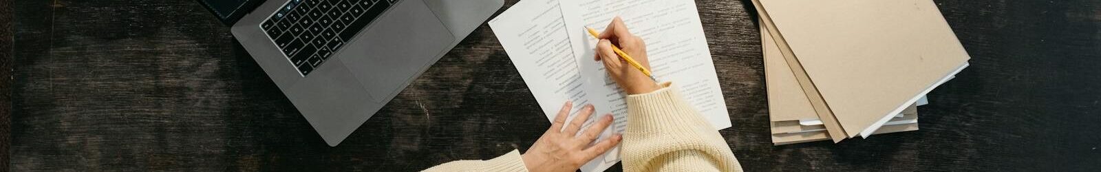 Overhead view of woman writing at desk with laptop, papers, and books.